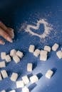 Top view of sugar cubes and a heart drawn in sugar on a blue surface with a hand nearby.